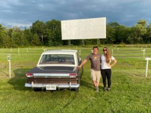 A man and a woman stand in a field at a drive-in theatre next to an antique car