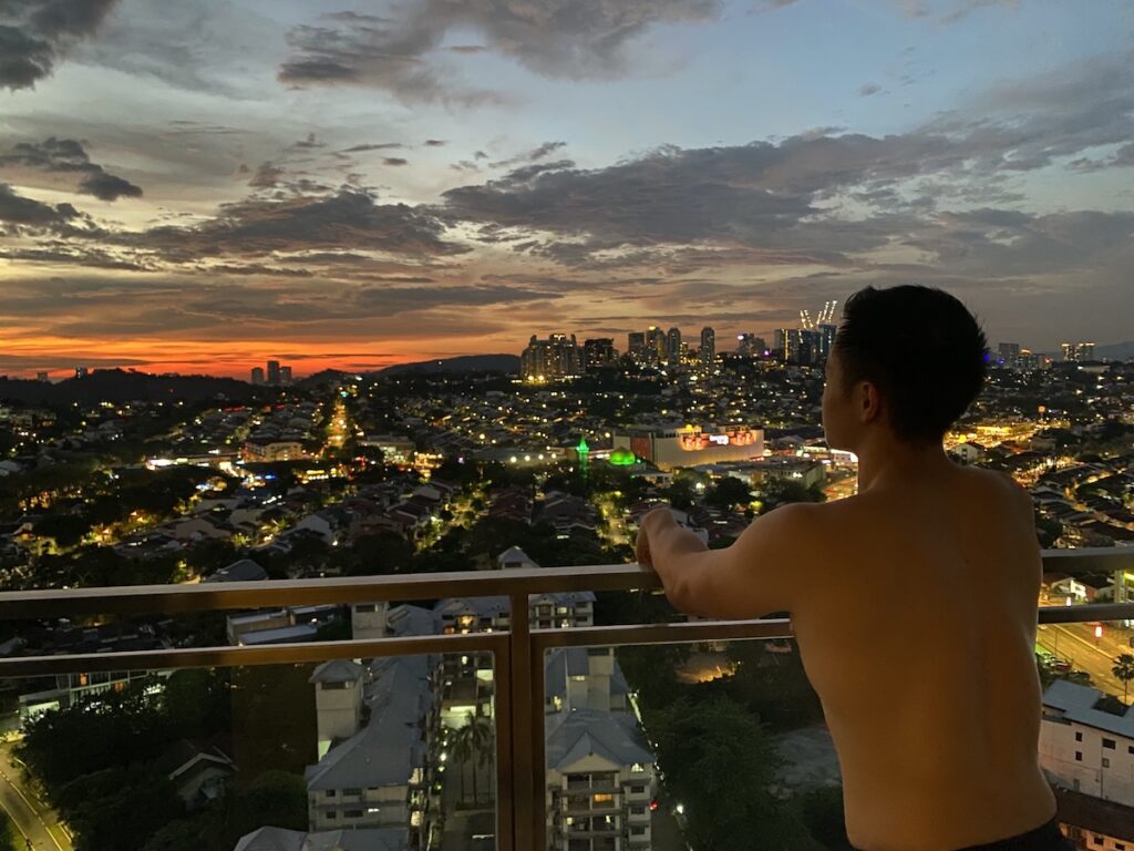 shirtless man leans on balcony overlooking sunset in city